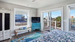 Un dormitorio con una cama grande, televisor, cómoda y ventana con vistas a edificios y un balcón.