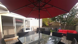 Une terrasse extérieure avec une table en verre et des chaises en osier, un parasol rouge et un canapé rouge en arrière-plan.