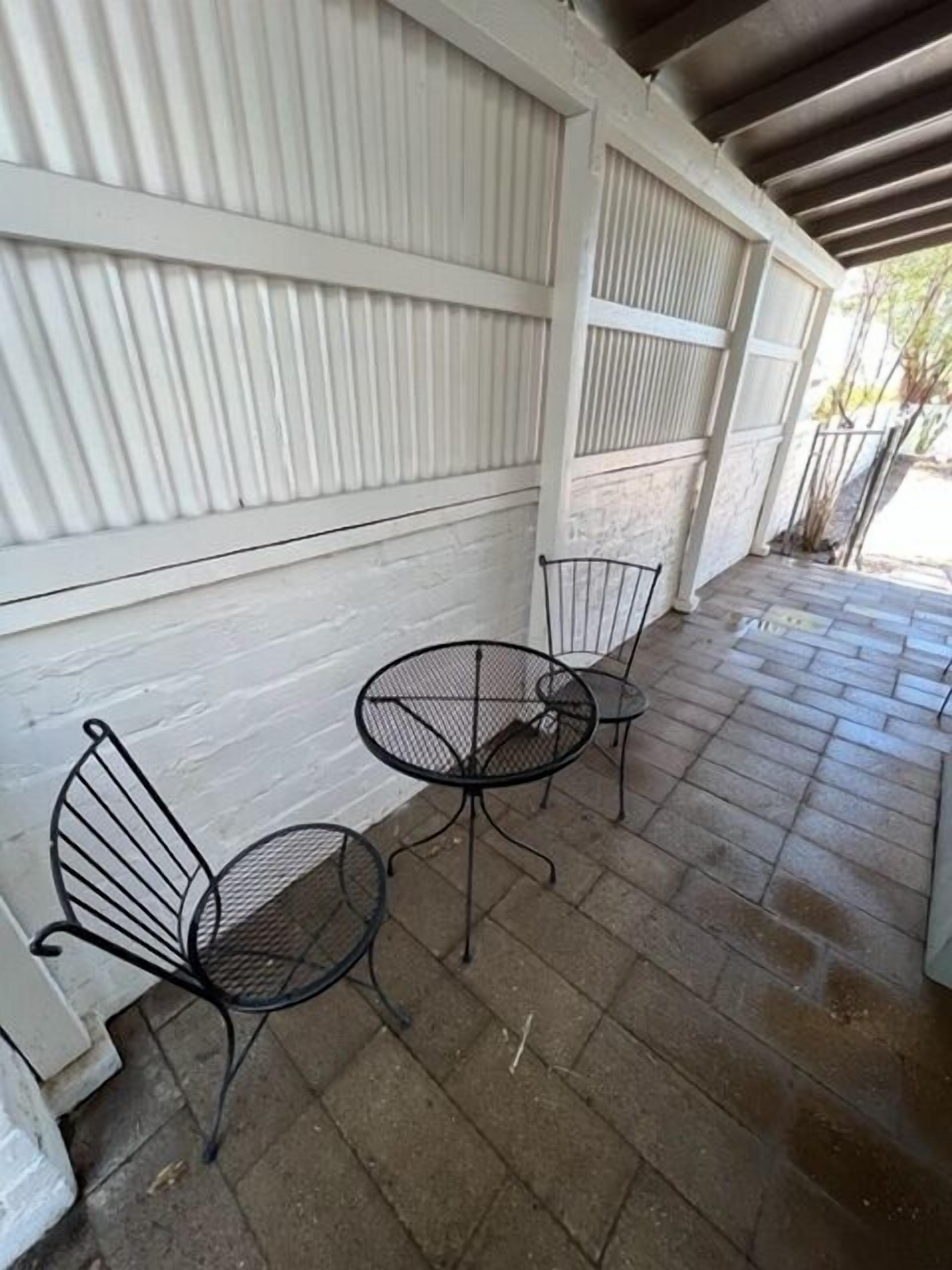 Een overdekt terras met twee metalen tafels en stoelen, een witte muur met verticale lamellen en een betegelde vloer.