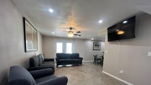 A living room with two sofas, a ceiling fan, a flat-screen TV mounted on the wall, and a dining area with chairs and a table.