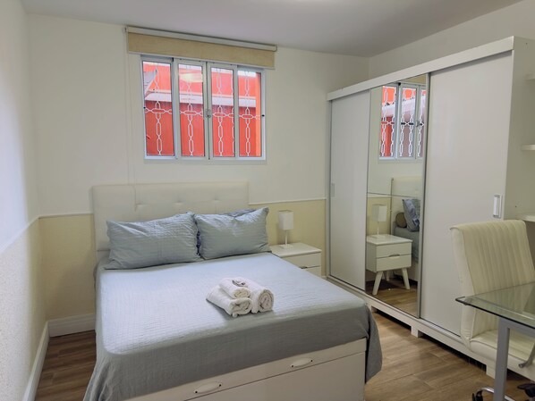 A bedroom with a bed, a bedside table, a mirror, a chair, and a window with a view of a balcony.