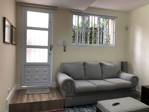 A living room with a grey sofa, a small table, a door with glass panels, and a window with a metal grille.