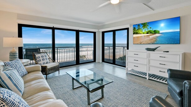 A spacious living room with a large window overlooking a beach, a glass coffee table, and a white dresser.