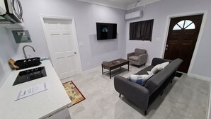 A modern living room with a sofa, a coffee table, and a TV. There is a door, a window with curtains, and an air conditioning unit on the wall. - Yamabeach Stays 3 (Nassau)