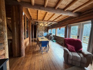 Interior de una cabaña de troncos con una mesa de comedor de madera, sillas azules, un sofá rojo, paredes de piedra y ventanas grandes.