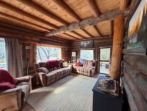 Interior de una cabaña de troncos con un sofá, una chimenea y un televisor montado.