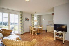 A living room with a TV, a dining table, and chairs. There is a window with curtains and a view of the outside.