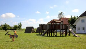 Ein Spielplatz mit einer Holzrutsche und Klettergerüsten, umgeben von einer Wiese und einem Gebäude mit rotem Dach.