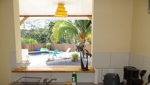 Una cocina con un lavamanos, un tostador y una ventana que da a un patio trasero con piscina y sillas de descanso.