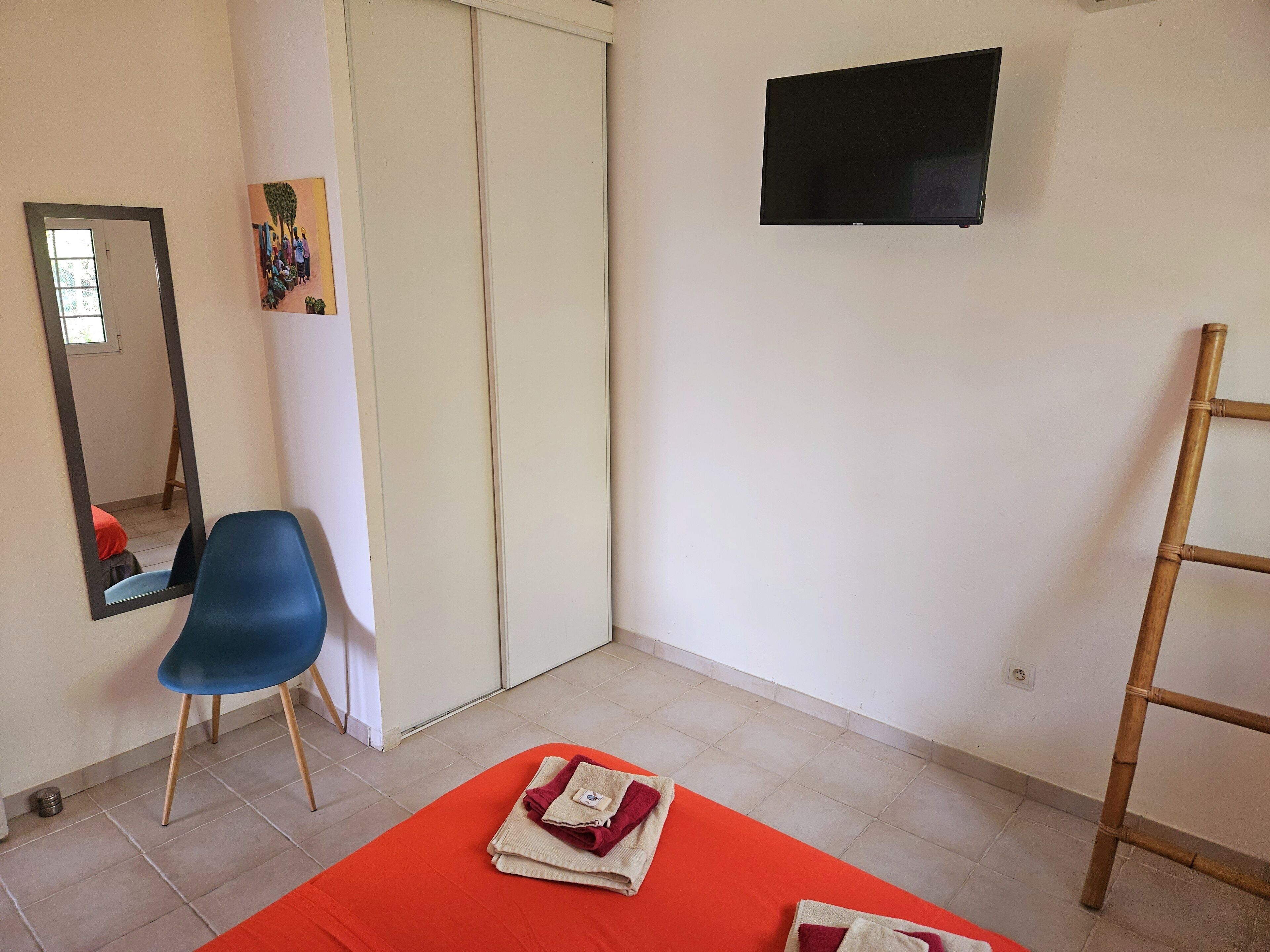 A room with a blue chair, a red bedspread, a wooden ladder, and a TV on the wall.