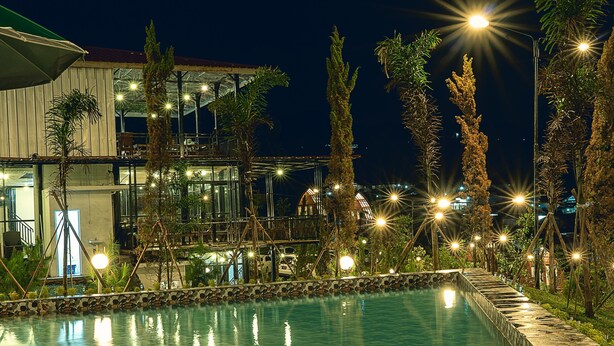 A nighttime view of a swimming pool with surrounding palm trees and a building with multiple levels and balconies. - Miracle Signature Hills (Ciater)