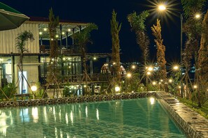 Vista noturna de uma piscina cercada por palmeiras e um edifício com vários andares e varandas.