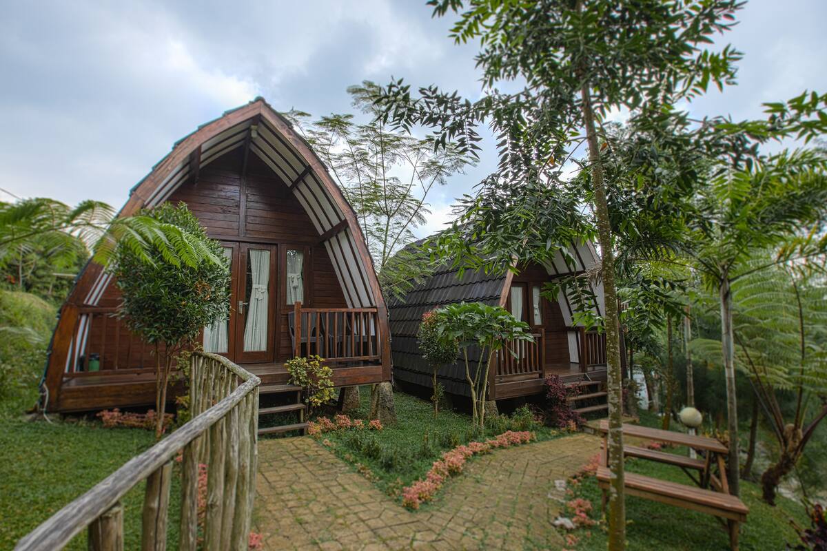 Two wooden cabins with pitched roofs surrounded by lush greenery and a wooden pathway.