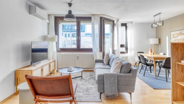 Ein modernes Wohnzimmer mit einer Couch, einem Couchtisch und einem Fernseher, ein Essbereich mit Stühlen und einem Tisch sowie große Fenster mit Blick auf Gebäude.