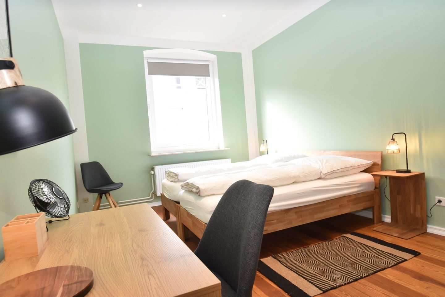A bedroom with a bed, a desk, a chair, a standing lamp, and a window.