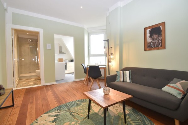 A living room with a sofa, a coffee table, and a dining area with a chair and table. There is a kitchen visible in the background.