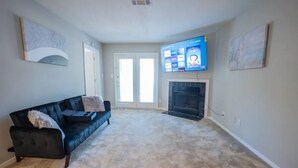 A living room with a black leather sofa, a flat-screen TV mounted above a fireplace, and a beige carpet. - Cozy 2-Bedroom Suite Getaway (Sandy Springs)