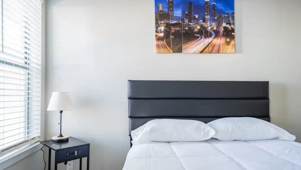 A hotel room with a bed, a nightstand with a lamp, and a cityscape painting on the wall. - Cozy 2-Bedroom Suite Getaway (Sandy Springs)