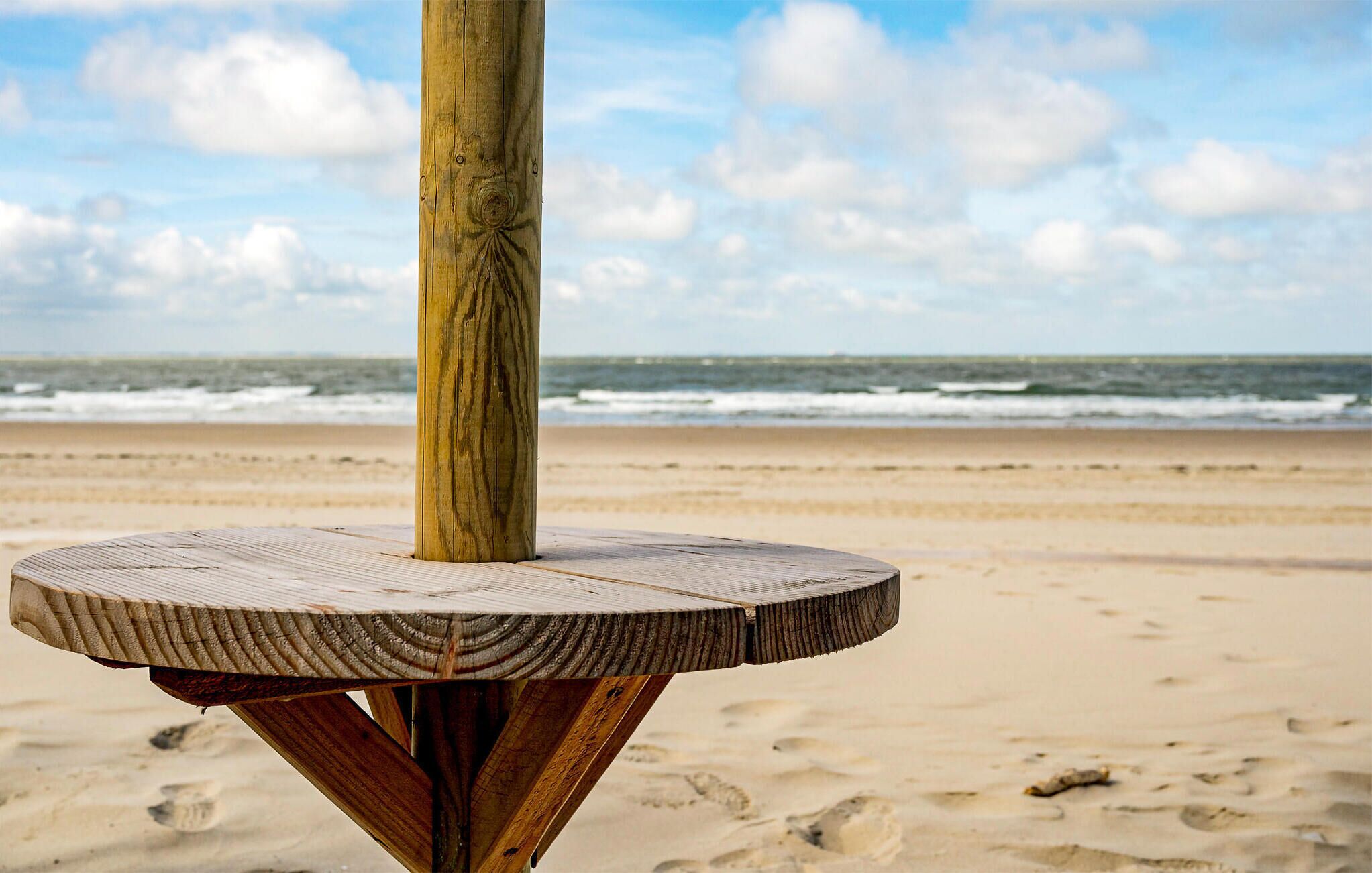 Une table en bois, dotée d’un plateau rond et d’un pied central, est placée sur une plage de sable, avec l’océan en arrière-plan.