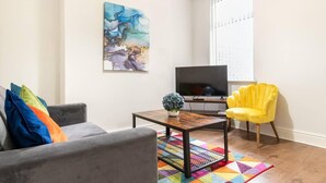 A living room with a gray sofa, a yellow armchair, a coffee table, a flat-screen TV, and a colorful geometric rug.