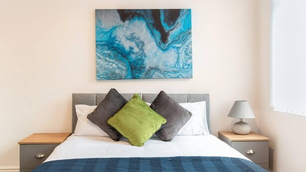 A neatly made bed with a blue blanket, a marble artwork above it, and a bedside lamp on a wooden nightstand.