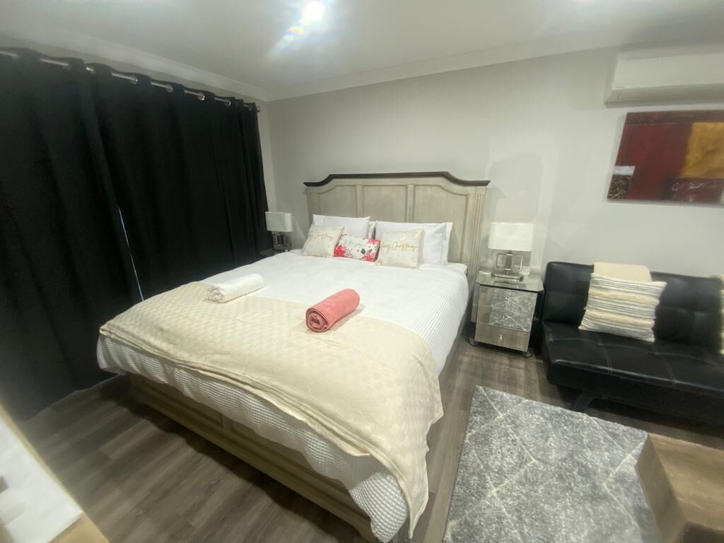 Un lit bien fait, avec une couette blanche et une serviette rose, un canapé en cuir noir, une petite table de chevet avec une lampe et un coussin décoratif.