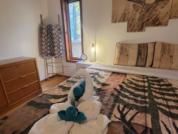 A bedroom with a bed, a wooden dresser, a window with blinds, a nightstand with a lamp, and a wall with a painting.