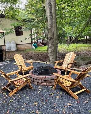 Relax in a serene outdoor seating area surrounded by trees and a wooden fence, featuring a fire pit and Adirondack chairs.