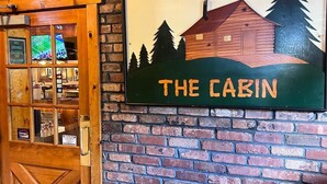 A rustic wooden door with a glass panel, a wall-mounted sign reading "THE CABIN" with a cabin illustration, and a brick wall. - Cabin for up to 10 People ! (Tobyhanna)