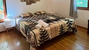 A bedroom with a wooden ceiling, a bed with a leaf-patterned bedspread, a nightstand with a clock, and a window with a view of trees. - Cabin for up to 10 People ! (Tobyhanna)