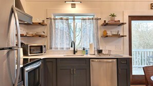 Una cocina moderna con armarios oscuros, microondas, lavavajillas y una ventana con cortinas traslúcidas.