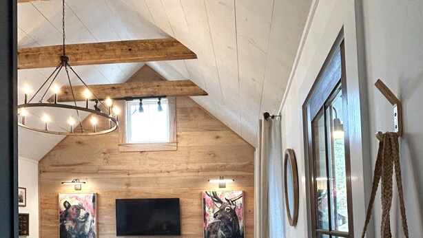 Una habitación estilo loft acogedora, con techo de madera, zona de comedor con mesa y sillas, sofá, televisor montado en la pared y un ventanal con cortinas.