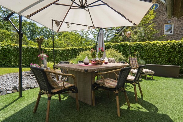 An outdoor patio with a table and chairs, surrounded by greenery and a brick building.