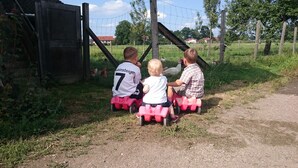 Three children on pink toy cars facing a wooden gate. - Ferienwohnung Nummer 2 im Zweiten Stock (Übersee)