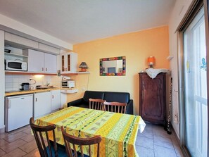 Cozinha compacta com armários brancos, micro-ondas e torradeira. Há uma mesa de jantar com toalha amarela e duas cadeiras. Um armário com toalha de estampa de leopardo é visível, e um espelho pendurado na parede.