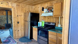 A cabin kitchen with wooden walls, a stainless steel refrigerator, a black stove, and a stone fireplace. - Historic Log Cabin in Greer, AZ.  Located near Molly Butler's & Sunrise Ski Area (Greer)