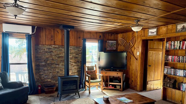 A cozy cabin interior with a stone wall, a fireplace, a television, a bookshelf, and a wooden ceiling. - Historic Log Cabin in Greer, AZ.  Located near Molly Butler's & Sunrise Ski Area (Greer)