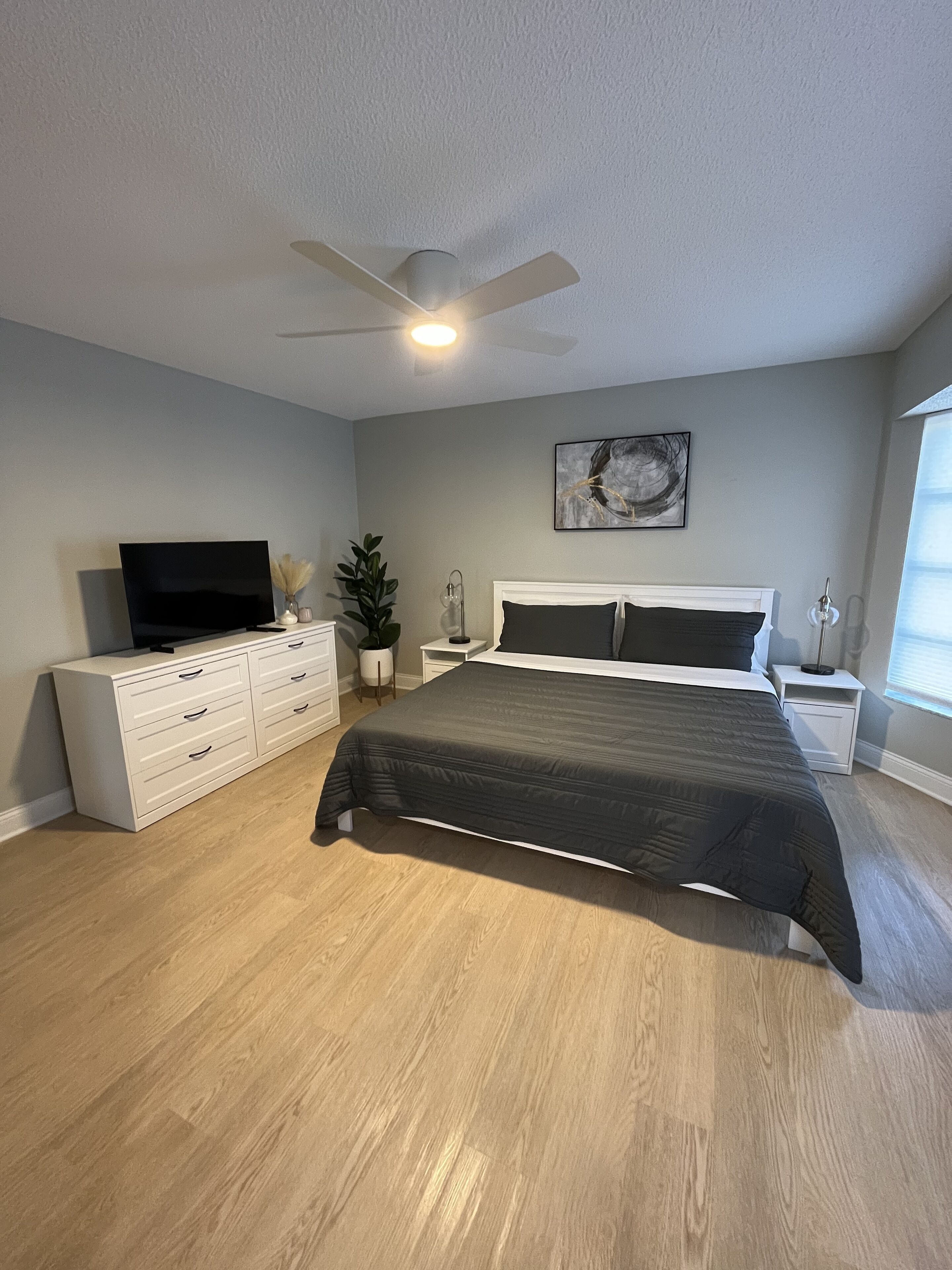 A bedroom with a bed, dresser, TV, ceiling fan, and artwork on the wall.