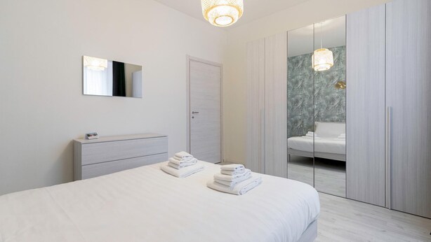 Un dormitorio moderno con cama, mesita de noche, espejo, armario y baño visible a través de una puerta de cristal.