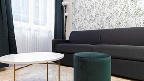 Un salón con un sofá gris oscuro, una mesa redonda blanca con patas doradas y un reposapiés verde.