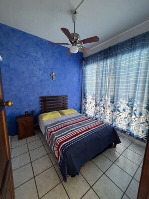 Une chambre à coucher avec un lit, une table de chevet, un ventilateur de plafond et une fenêtre avec des rideaux.