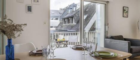 Un coin repas avec une table dressée pour quatre personnes, un vase bleu contenant des fleurs séchées et une vue sur un balcon avec une balustrade et un bâtiment en arrière-plan.