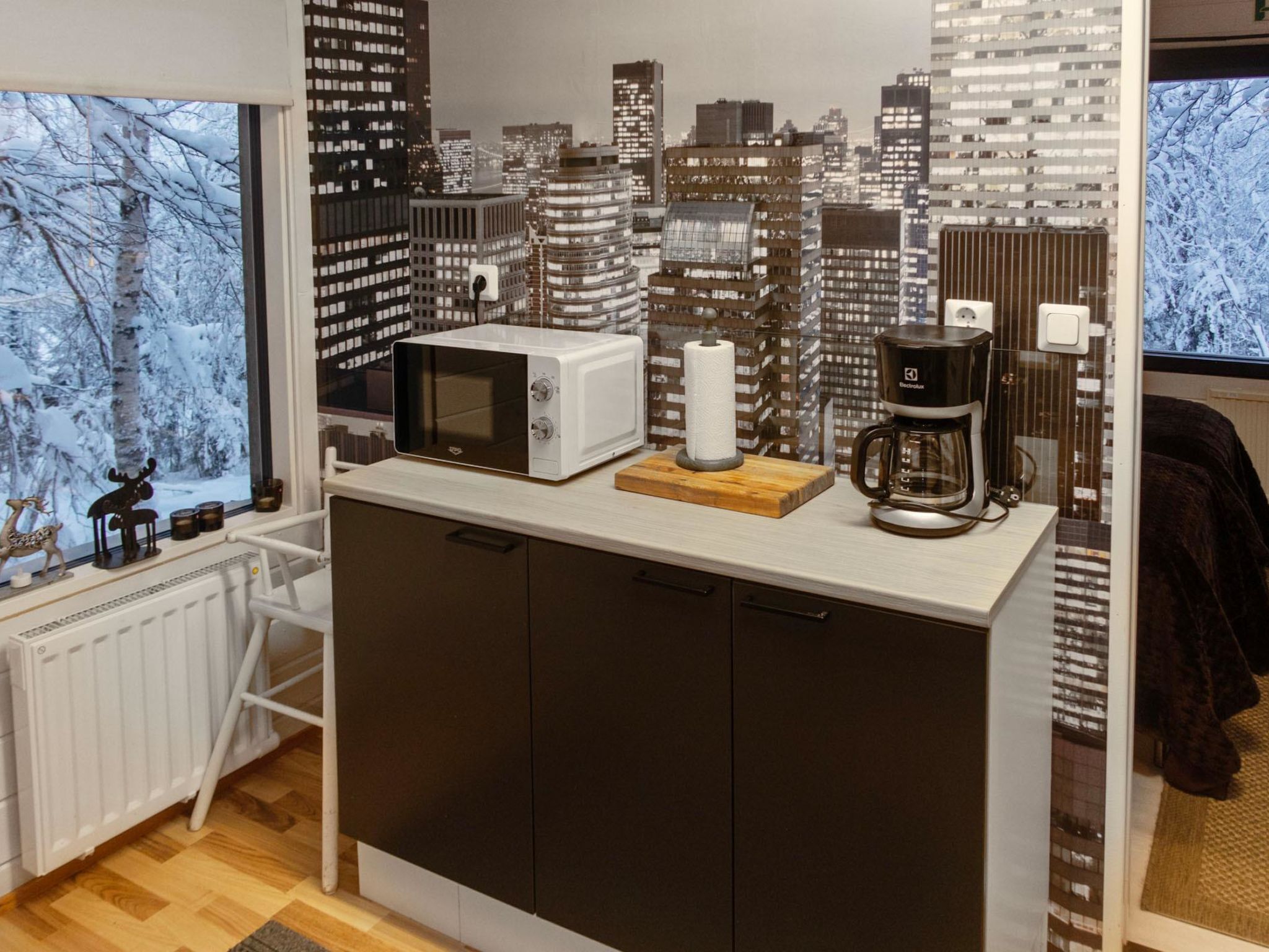 A kitchen with a view of a snowy forest, a microwave, a coffee maker, and a roll of paper towels on the counter.