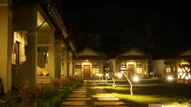 A pathway leading to a row of buildings with illuminated windows and doors, surrounded by greenery. - Breathing Earth (Diamond Harbour)