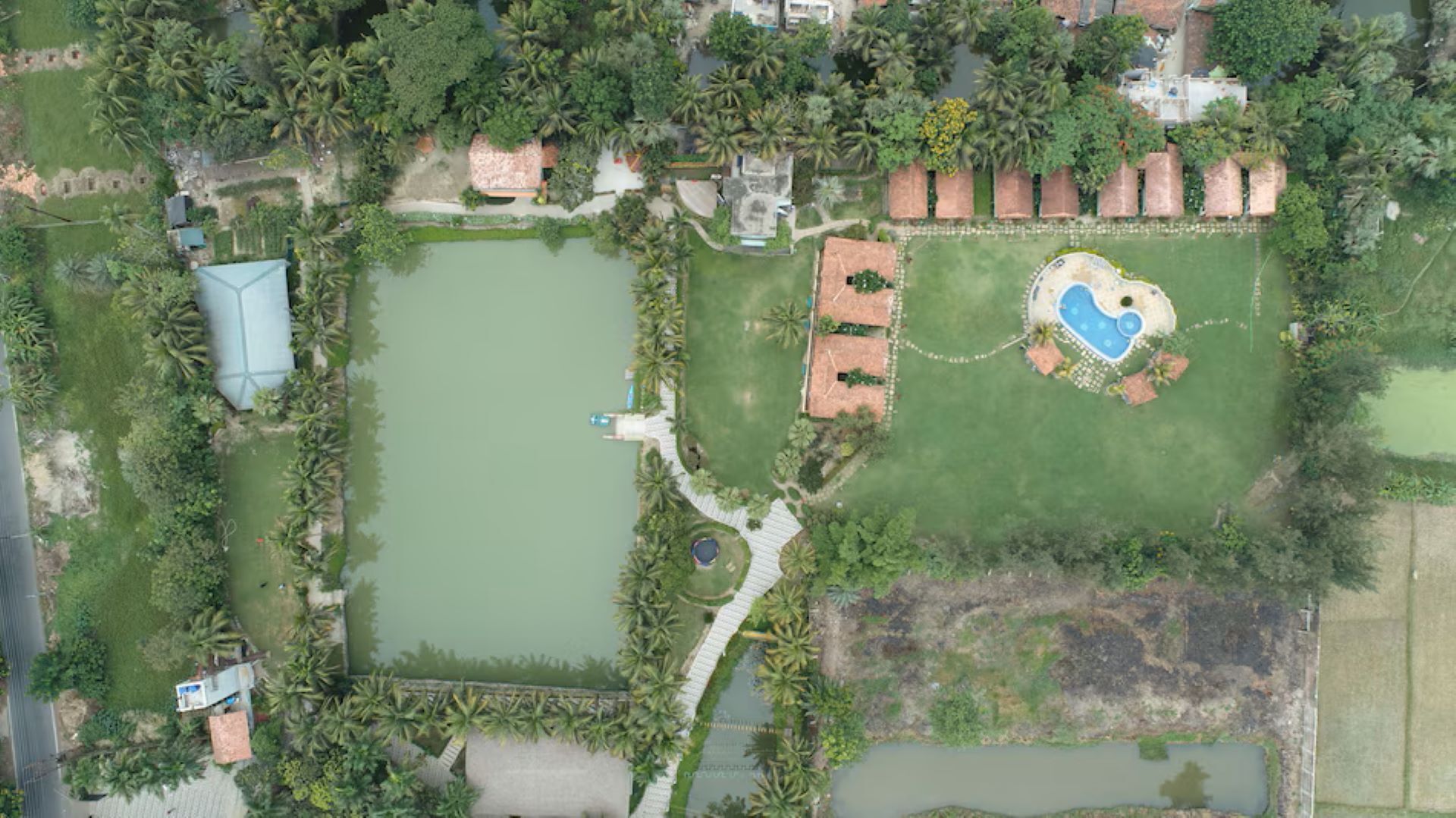 An aerial view of a property with a large central pond, a swimming pool with a heart-shaped design, and surrounding greenery.