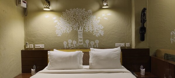 Habitación de hotel con cama, dos almohadas, mesita de noche, lámpara de pared y un mural decorativo con un árbol.