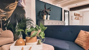 Een gezellige woonkamer met een blauwe bank, een roze pouf en een witte bijzettafel met een vaas en planten.