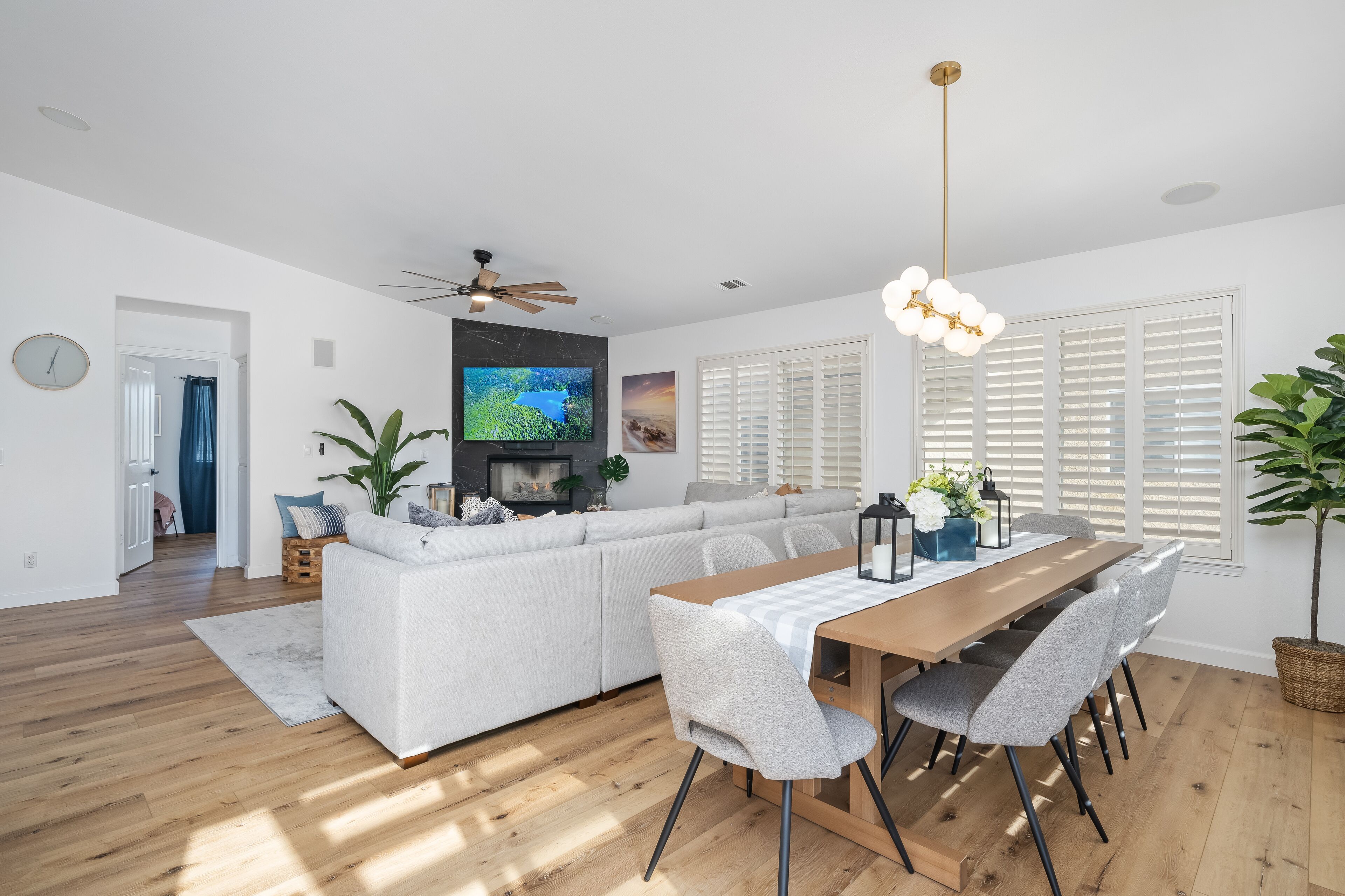 Een moderne woonkamer met een bank, een eettafel en stoelen. Er is een tv aan de muur bevestigd, een plafondventilator en een groot raam met zonwering.