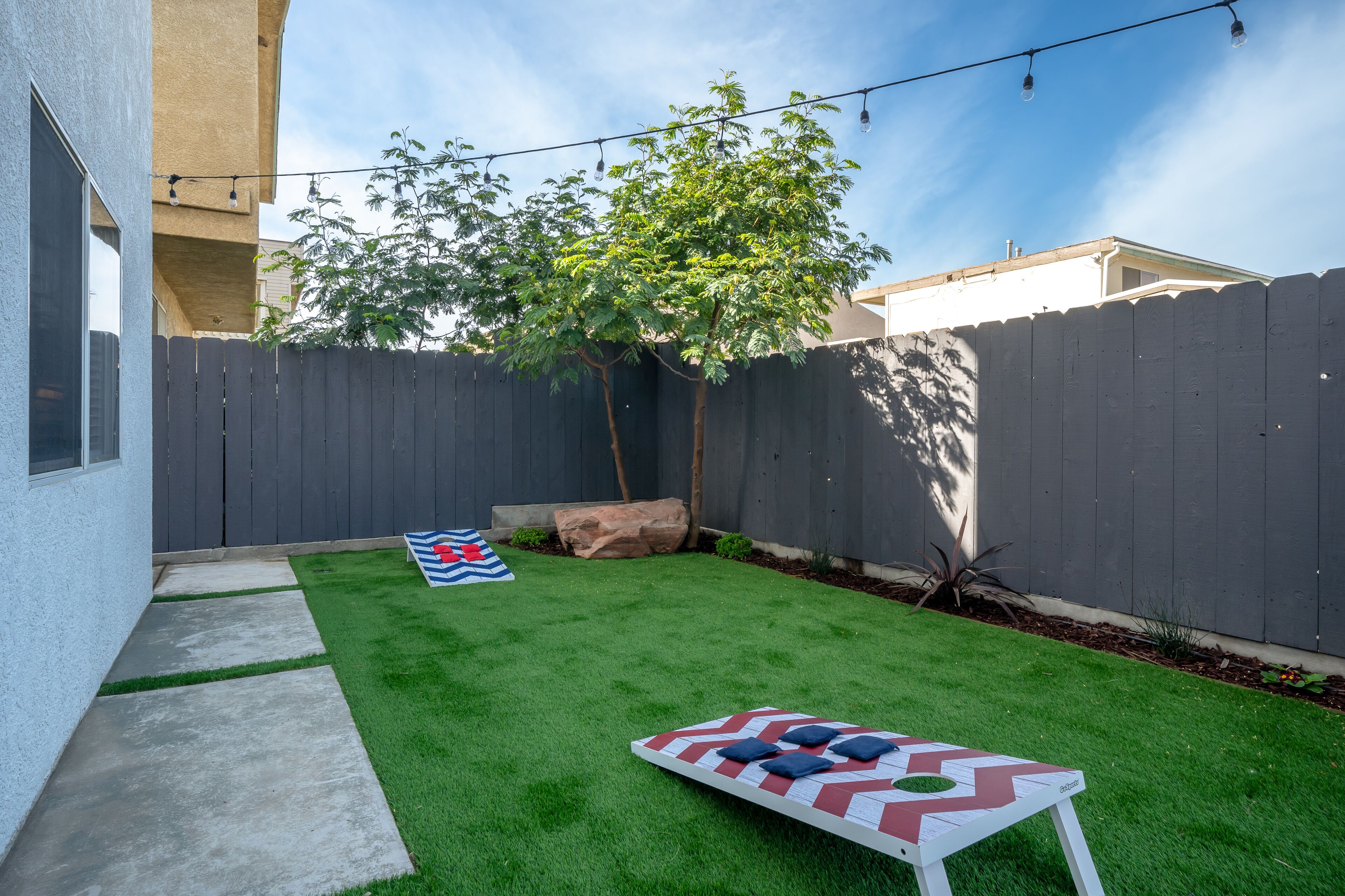 A backyard with artificial grass, a wooden fence, a tree, and a table with a checkered tablecloth and blue cushions.