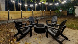 Un área de estar al aire libre con cuatro sillas Adirondack y un pozo de fuego circular, rodeada por una cerca de madera e iluminada con luces colgantes.